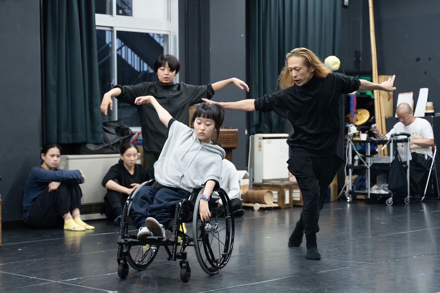 車椅子に乗り片手を挙げて踊る和合由依と、そのそばで両手を広げて踊る森山開次、後ろにはキャストが数人写っている写真