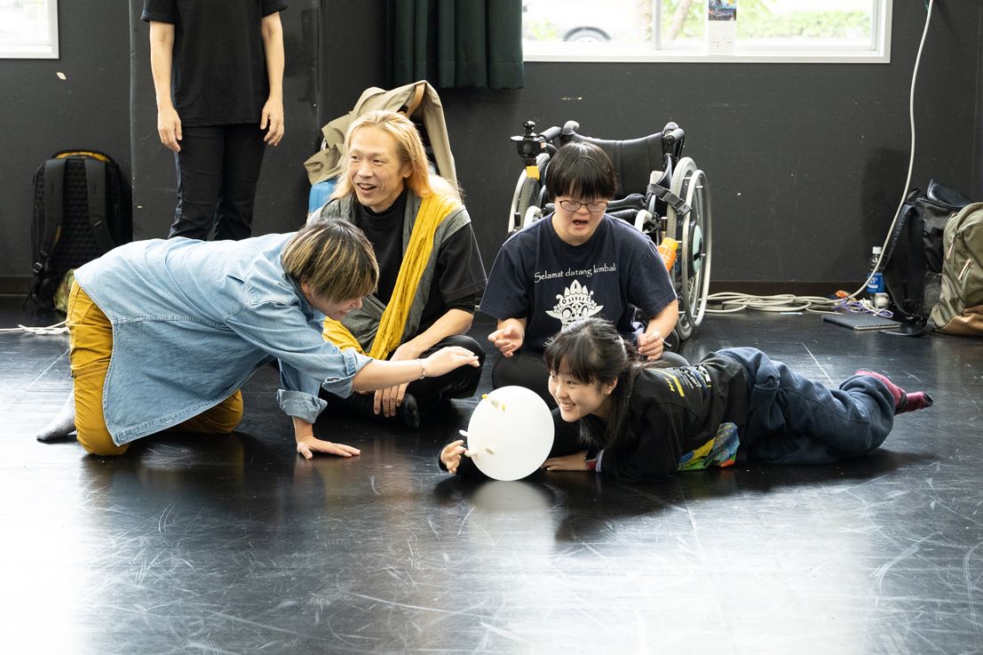 稽古場の床に座った4人が、一つの白い風船をキャッチしようとしている。